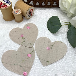 May include: Three heart-shaped fabric patches with pink embroidered flowers and vines. The patches are made of a natural linen fabric and are perfect for adding a touch of handmade charm to any project.