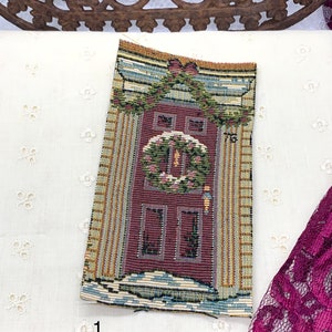 May include: A tapestry depicting a red door with a wreath and a window above it. The door is surrounded by a yellow and green frame. The tapestry is on a white background.