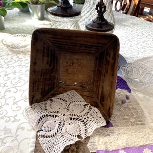 May include: A square, wooden, rustic basket with a white crocheted doily inside. The basket is brown with visible wood grain. Decorative glass jars with fleur-de-lis lids are in the background. A vintage-themed display.