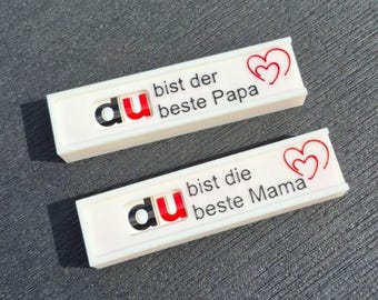 May include: Two white rectangular badges with black and red text. One says "du bist der beste Papa" with a red heart. The other says "du bist die beste Mama" with a red heart. The badges are on a dark gray surface.