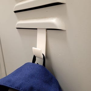 Puede incluir: Un gancho de plástico blanco montado en una puerta de casillero de metal con una mochila azul colgando de él.