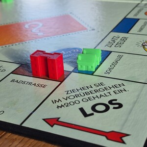 Könnte beinhalten: Nahaufnahme eines Monopoly-Spielbretts mit einem roten und grünen Haus auf dem Feld "Los". Der Text "ZIEHEN SIE IM VORÜBERGEHEN M200 GEHALT EIN. LOS" ist auf dem Brett gedruckt.