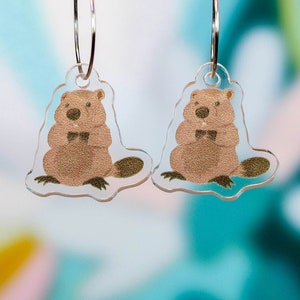 May include: A pair of clear acrylic earrings featuring a brown beaver illustration on each. The beavers are wearing a bow tie and have a gold glitter texture.