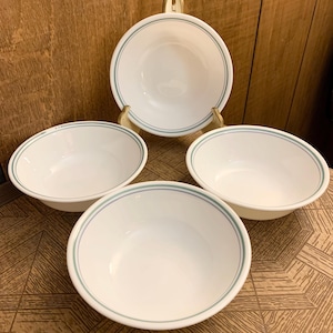 Puede incluir: Cuatro cuencos de cerámica blanca con un borde azul claro. Un cuenco está elevado sobre un soporte dorado. Los cuencos están dispuestos sobre una superficie de madera. Los cuencos están vacíos y listos para usar.