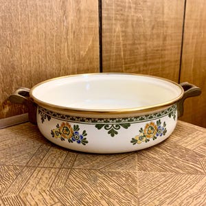 May include: Cream-colored enamelware bowl with floral design. The bowl has a gold rim and handles. The floral pattern includes blue, yellow, and orange flowers with green leaves. The bowl sits on a wooden surface.