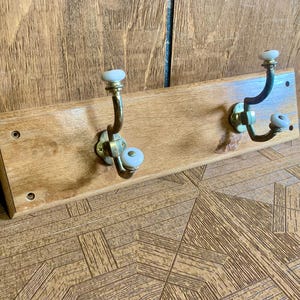 Op de afbeelding: Houten kapstok met twee messing haken. De haken hebben witte keramische knoppen. De houten plank is lichtbruin met zichtbare houtnerf.
