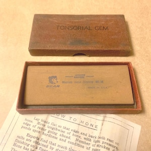 May include: A vintage brown rectangular box labeled "TONSORIAL GEM" with a smaller box inside. The inner box contains a sharpening stone with the text "BEAR" and "Medium India Oilstone MS 34". A paper with instructions is also visible.