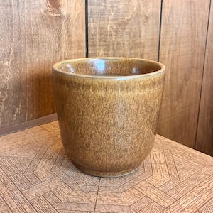 May include: A ceramic mug with a speckled, mottled brown glaze. The vessel features a cylindrical shape with a slightly tapered base and a smooth, rounded rim. The finish displays variations in color intensity, ranging from light tan to deep earth tones.