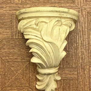 Può includere: Una mensola da parete color crema con finiture anticate e dettagli scuri. L'oggetto presenta un piano superiore semicircolare e un motivo decorativo intagliato a forma di foglie che si restringe verso il basso fino a una punta.