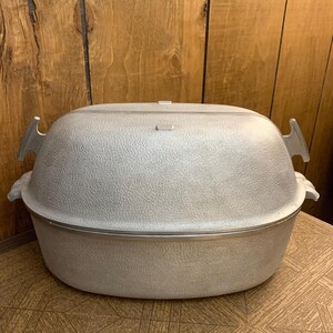 May include: A light gray oval-shaped roaster with a textured surface and a matching lid. The roaster has two handles on the sides and a small handle on the lid. The roaster is sitting on a patterned surface.