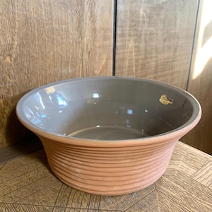 May include: A terracotta ceramic bowl with a ridged exterior and a smooth, grey interior. The bowl has a wide, slightly flared rim. A small, gold-coloured sticker is visible on the rim. The bowl is on a wooden surface.