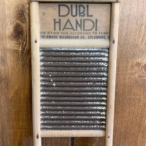 Op de afbeelding: Een vintage houten wasbord met de tekst "DUBL HANDI" en "COLUMBUS WASHBOARD CO.". Het wasbord heeft een gegolfd metalen schrobvlak en een houten frame. Een klassiek wasmiddel.