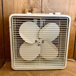 May include: A vintage, off-white box fan with four blades, encased in a white metal grill. The fan has a square shape and a handle on top. The blades are visible behind the protective grill. The fan is plugged into a wall outlet.