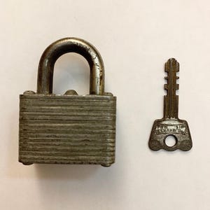 May include: A vintage, weathered padlock with a matching key. The padlock is rectangular with horizontal ridges and a curved shackle. The key is a small, ornate design with a circular hole. The key has the word "MASTER" on it.