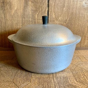 Op de afbeelding: Een vintage, zilverkleurige kookpot met een deksel. De pot heeft een getextureerd oppervlak en een zwarte knop bovenop het deksel. De pot heeft twee handvatten aan de zijkanten. De pot staat op een houten oppervlak.
