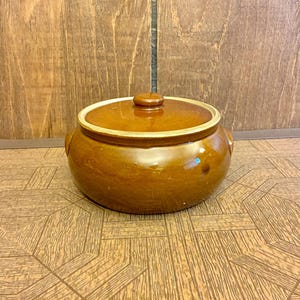 以下が含まれることがあります： 蓋付きの茶色の釉薬をかけた丸い陶器製のキャセロール皿。鍋には2つの統合されたサイドハンドルと、蓋の中央に小さなつまみが付いています。蓋の縁と鍋の上端は明るいベージュ色で仕上げられています。
