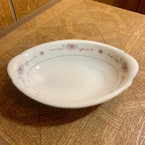 Puede incluir: Un plato ovalado blanco para servir con un diseño floral en rosa, azul y verde. El plato tiene un borde festoneado y un borde dorado.