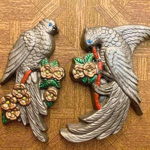 May include: Two silver-colored bird wall decorations. Each bird has blue eyes and is perched on a branch with flowers. The flowers are in shades of brown, green, and yellow. The background is a brown, patterned surface.