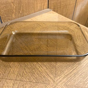 Puede incluir: Fuente de horno rectangular de vidrio color ámbar con esquinas redondeadas y asas. La fuente tiene un borde oscuro y está vacía. La fuente está sobre una superficie de madera.