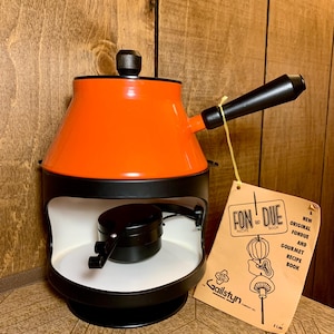 Puede incluir: Una olla de fondue naranja y negra con un asa y una tapa negras. La olla tiene un interior blanco y se asienta sobre una base negra. Un libro de recetas con el título "Fondue" cuelga de la olla.
