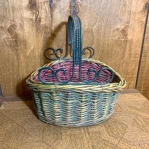 May include: Oval-shaped woven basket with a decorative metal handle and accents. The basket features a mix of pastel colors, including pink, green, and yellow. The metal handle is dark green with decorative swirls.