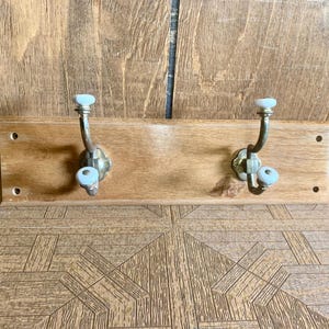 May include: Wooden wall-mounted coat rack with two metal hooks. The hooks have a decorative floral base and a light blue ceramic knob. The wooden board is rectangular and has a natural wood finish.