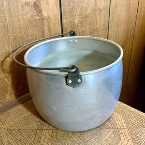 Puede incluir: Una olla de aluminio vintage con un cuerpo curvo y un asa de metal. La olla tiene un acabado plateado y está diseñada con un asa resistente para facilitar su transporte. La olla probablemente se utiliza para cocinar o almacenar.