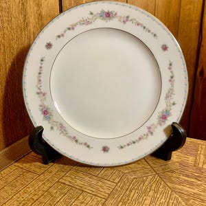 Puede incluir: Un plato de cena de cerámica blanca con un delicado patrón floral en tonos de rosa, azul y verde. El plato tiene un borde dorado y un sutil acento dorado en el centro.