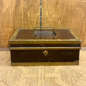 May include: A rectangular metal cash box with a dark brown finish and brass-colored trim. The box features a central keyhole on the front panel and a collapsible metal handle mounted on the top lid. The surface shows signs of wear and scratches.
