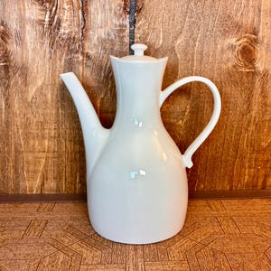 May include: A white porcelain coffee pot with a smooth, curved body, a tall neck, a long angled spout, and a matching lid with a small knob handle. The vessel features a wide base that tapers upward and a large, arched side handle.