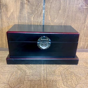 Pode incluir: Uma caixa de madeira preta com formato retangular e fecho de metal decorativo. A caixa tem uma borda vermelha e assenta sobre uma base correspondente. O fundo mostra o grão da madeira.