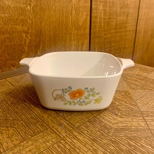 May include: White ceramic baking dish with a floral design featuring orange, blue, and green flowers. The dish has two handles on the sides.