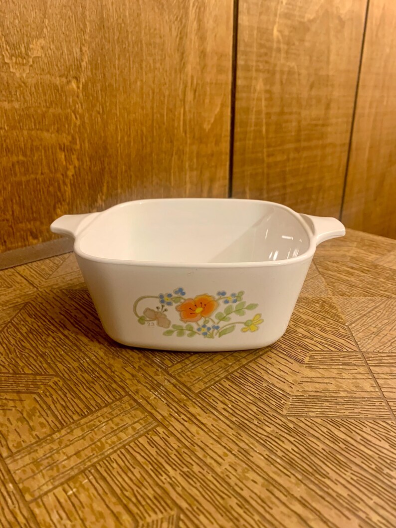 May include: White ceramic baking dish with a floral design featuring orange, blue, and green flowers. The dish has two handles on the sides.
