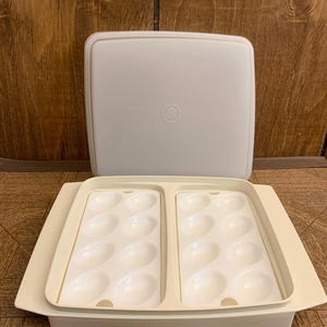 May include: A vintage, off-white plastic egg container with a matching lid. The container has two trays, each with 10 egg-shaped indentations. The lid is square with a circular indentation in the center. The container is open, revealing the egg holders.