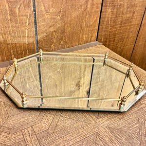 Könnte beinhalten: Ein sechseckiges Spiegeltablett mit einer polierten goldfarbenen Metallreling. Das Tablett hat einen reflektierenden Glasboden und wird an jeder Ecke von kleinen, dekorativen Metallpfosten gestützt, was eine flache Struktur zur Präsentation ergibt.