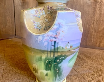 May include: A vintage Japanese porcelain vase featuring a hand-painted landscape scene. The vase is decorated with a light blue, purple, and green landscape with a white house and trees. The vase has a gold rim and a scalloped top.