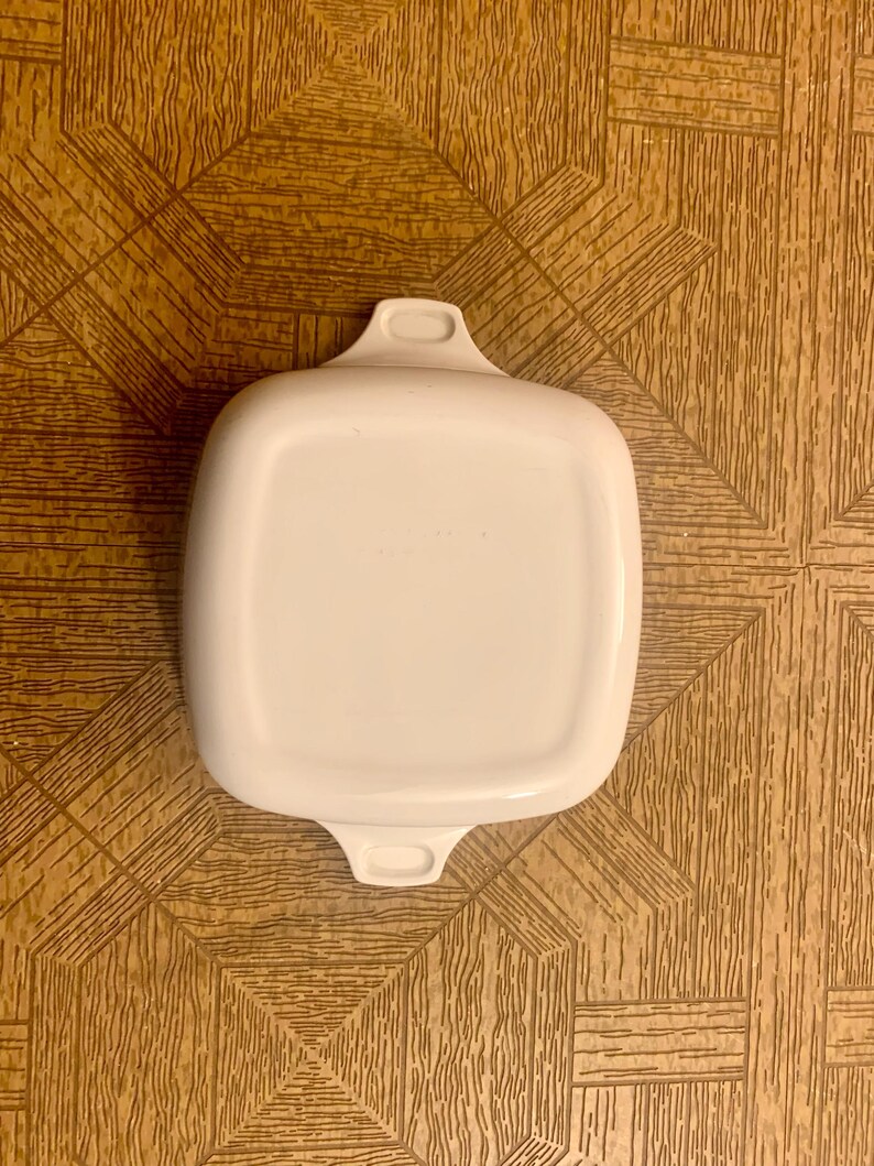 May include: White square baking dish with two handles. The dish is made of ceramic and has a smooth, glossy finish.