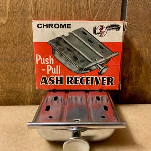 May include: Chrome push-pull ash receiver with a white knob. The product is in its original box, which is red and white with the words "Chrome" and "Push-Pull Ash Receiver". The box also has a vintage Hollywood logo.