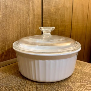 Op de afbeelding: Een witte, geribbelde ovenschaal met een doorzichtig glazen deksel en een klein handvat. De schaal staat op een houten oppervlak. Deze schaal wordt waarschijnlijk gebruikt voor het bakken en serveren van voedsel. Ongeveer 20 cm in diameter.
