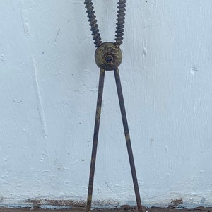May include: An antique metal kitchen utensil, possibly a pair of tongs or a similar tool. The handle is a coiled spring, and the tongs have a rusty, aged appearance. The background is a white wall.
