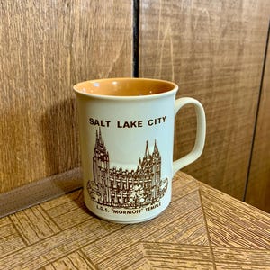 Puede incluir: Taza de cerámica color crema con una ilustración marrón de un edificio y el texto "SALT LAKE CITY" y "L.D.S. 'MORMON' TEMPLE". El interior de la taza es naranja. Tiene un asa curva.