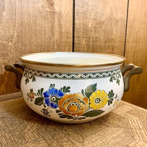May include: A white enamelware bowl with a floral design. The bowl features blue, orange, and yellow flowers with green leaves. It has a gold rim and two brown handles. The bowl is sitting on a wooden surface.