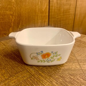 May include: White square baking dish with a floral design featuring orange, blue, and yellow flowers. The dish has two handles on the sides.