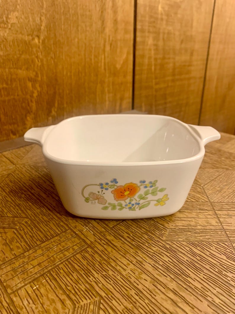 May include: White square baking dish with a floral design featuring orange, blue, and yellow flowers. The dish has two handles on the sides.