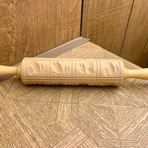May include: A light-colored wooden rolling pin with carved bird designs. The rolling pin has two handles and a patterned rolling surface. The wood has a natural finish, and the design is carved into the wood.