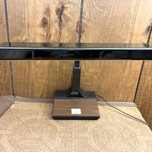 May include: A black desk lamp with a long, rectangular horizontal light head and a vertical support post. The base features a wood-grain finish with two white rectangular power switches centered on the front surface and a black power cord.