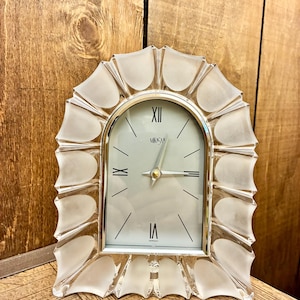 May include: An arched mantel clock featuring a clear and frosted glass frame with a scalloped edge. The clock face is white with silver-tone hands, thin black hour markers, and the Roman numerals XII, VI, and IX. The brand name Mikasa is printed.