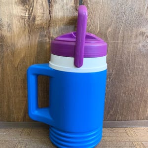 Puede incluir: Un termo azul y morado con una banda blanca y un asa morada. El termo tiene un asa grande y una tapa de rosca. El termo está diseñado para mantener las bebidas frías. El termo es de plástico.