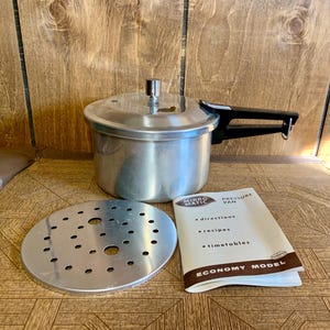 Könnte beinhalten: Ein Vintage Mirro-Matic Schnellkochtopf mit silbernem Finish und schwarzem Griff steht auf einer Holzoberfläche. Enthalten sind ein perforierter Metalleinsatz und eine Bedienungsanleitung. Das Heft trägt die Aufschrift "Pressure Pan" und "Economy Model".