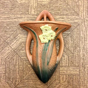 Puede incluir: Un florero de pared de cerámica con un acabado de terracota texturizado y un degradado verde oscuro en la base. El florero está adornado con un relieve floral de color amarillo pálido y dos asas laterales, rematado con una pestaña puntiaguda.